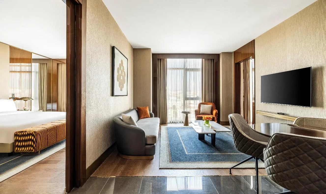 Suite living area: armchairs, coffee table and a city view through the window; the bedroom on the left